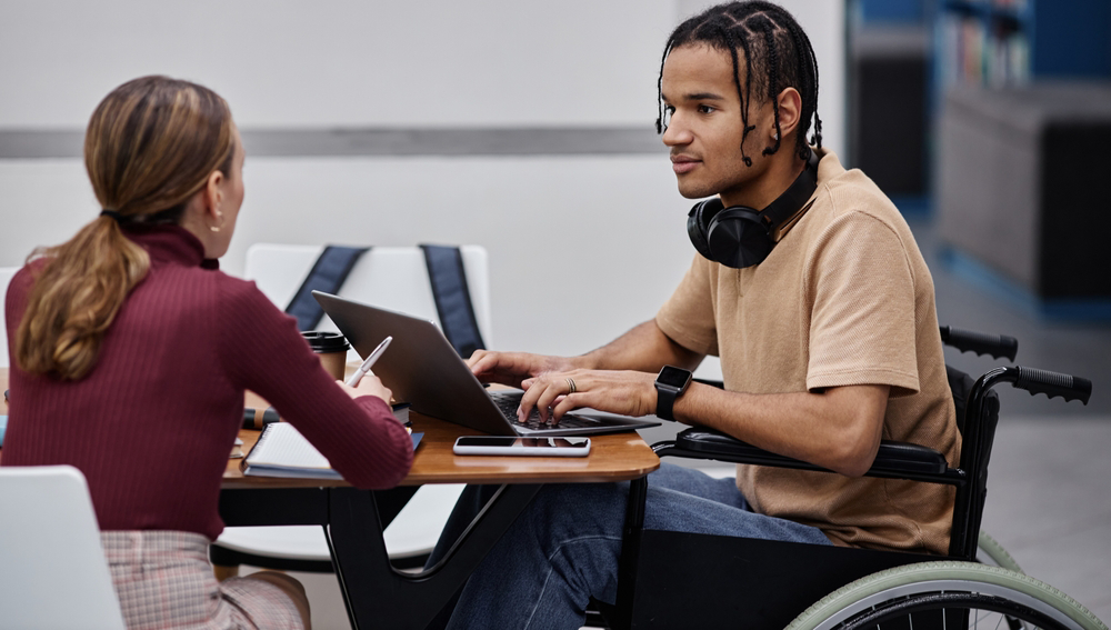 young male student in wheelchair in university library in conversation with young female student, both sat at table